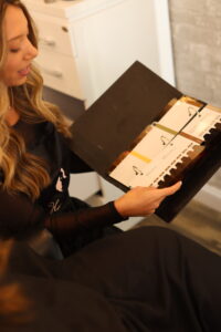 A woman with long blonde hair, wearing a black apron, looks at a color chart book. The book contains various hair color swatches. She appears to be in a salon setting, with drawers and a brick wall in the background.