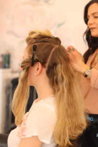 A woman with partially curled blonde hair sits in a salon chair. A stylist with long dark hair clips and styles her hair. The woman wears a white top, while the stylist wears a peach-colored blouse. The background shows salon equipment and a mural.