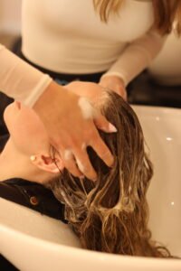 A person with long hair is reclining with their head in a white sink, while another person is washing their hair. The person washing the hair has their fingers lathered with shampoo and is massaging the scalp. Both individuals are wearing long-sleeve shirts.