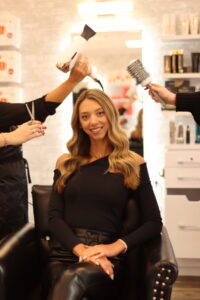 A woman with long, wavy hair sits in a salon chair, smiling. She is dressed in a black outfit. Two hairstylists work on her hair; one is holding a brush and the other a hairdryer. Shelves with hair products are visible in the background.