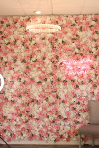 A lush, intricate flower wall adorned with pink and white roses serves as a vibrant backdrop. A circular chandelier hangs from the ceiling. A neon sign on the wall reads "This is where the magic happens." A light grey chair sits in the bottom right corner.