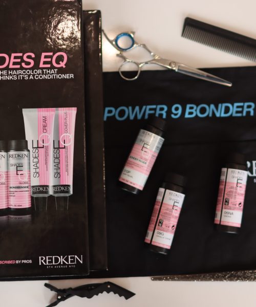 A display of Redken hair products includes a Shades EQ box containing hair color treatments, scissors, a comb, four small bottles, and an apron reading "POWFR 9 BONDER INSIDE." Brushes also lie nearby on a flat surface.